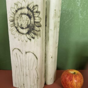 May include: A tall, off-white rectangular vase with a textured surface featuring a dark, etched sunflower design on the front. The vase has a vertical split down the side and is positioned next to a single, round, red and yellow apple.