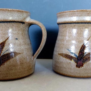 May include: Two handmade ceramic mugs. The mugs are a light tan colour with a dark blue band near the rim and a brown and blue leaf design. Each mug has a large handle.