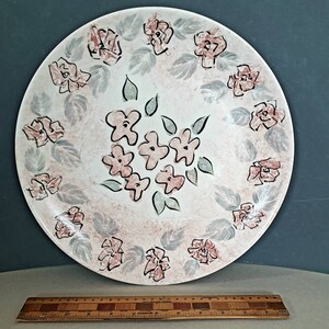 May include: A large, round, ceramic plate with a floral design. The plate has a white background with a pink and grey floral pattern. The flowers are in shades of pink and the leaves are grey and green. A wooden ruler is at the bottom.