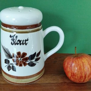 May include: A white ceramic flour shaker with a handle and a lid with holes. The word "Flour" is written in black script. The shaker is decorated with brown and black floral designs. An apple with red and yellow stripes sits next to the shaker.