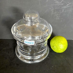May include: A clear glass jar with a rounded lid and a lime. The jar has a label that reads "Dartington Form Function". The lime is bright green and sits next to the jar.