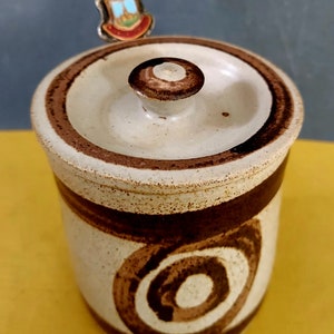 May include: A ceramic jar with a lid, featuring a speckled off-white body and brown spiral and band designs. The lid has a small knob and a brown rim. A small, decorative pin with a crest is on top.