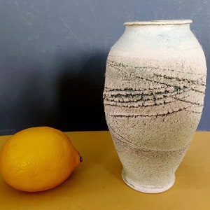 May include: A textured, off-white ceramic vase with a light blue top and a dark, linear pattern around the middle. A bright yellow lemon sits to the left of the vase. The vase is approximately 25 cm tall.