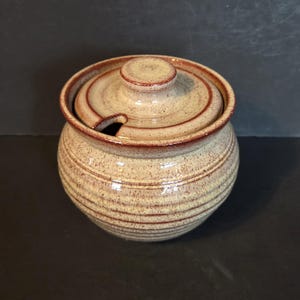 May include: A beige ceramic jar with a lid and a small opening for a spoon. The jar has brown horizontal stripes and a matching lid with a small knob. It is likely for storing sugar or other condiments. The jar is round.
