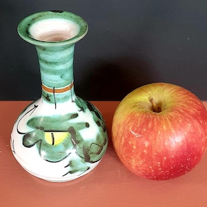 May include: A small, hand-painted ceramic vase with a white base and green and black abstract designs. The vase has a flared rim and a narrow neck. Beside the vase is a red and yellow apple.