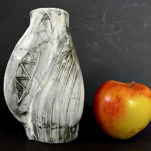May include: A handcrafted, grey and white ceramic vase with a textured surface and abstract designs. The vase is next to a red and yellow apple. The vase has a unique shape and is suitable for home decor.
