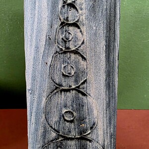 May include: A rectangular ceramic vase with a weathered, distressed finish in shades of blue, grey, and white. The front surface features a vertical arrangement of six embossed, concentric circular patterns that decrease in size towards the top.