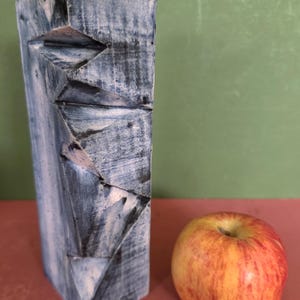 May include: A rectangular wooden block with a distressed blue and white painted finish featuring carved geometric facets along one side, positioned next to a round red and yellow apple for a clear sense of scale in this composition.