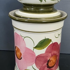 May include: A white ceramic canister with a lid, decorated with pink floral designs and green leaves. The canister has a cylindrical shape with a green trim and a matching lid. The floral pattern is hand-painted.
