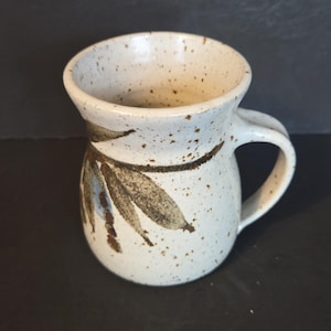 May include: A ceramic mug with a curved handle. The mug is off-white with brown leaf-like designs and brown speckles. The mug has a slightly flared rim. Suitable for hot or cold drinks, it measures approximately 10 cm in height.