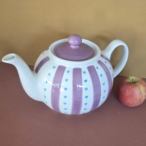 May include: A white ceramic teapot with a lilac lid and handle. The teapot features lilac stripes and small blue hearts. A red apple is placed beside the teapot.