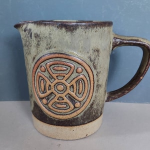 May include: A ceramic jug with a handle and a spout. The jug is mainly olive green with a brown rim and handle. A circular, embossed design in shades of brown and tan decorates the front. The base is a lighter tan colour.