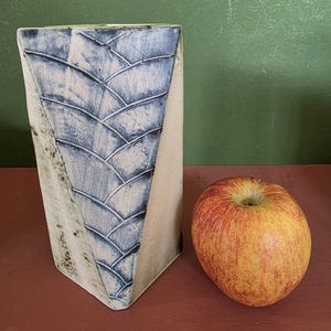 May include: A tall, rectangular ceramic vase with a white base and a prominent blue, textured, scale-like pattern on the front face. The vase is positioned next to a single, round, red and yellow apple, providing a clear sense of scale for the object.