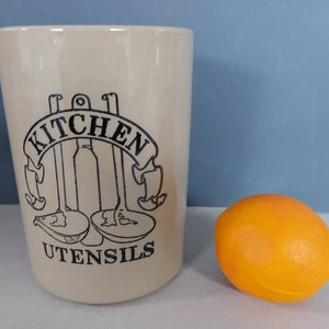 May include: A cream-coloured kitchen utensil holder with black lettering and illustrations. The text reads "KITCHEN UTENSILS" above drawings of kitchen tools. An orange fruit sits to the right of the holder.