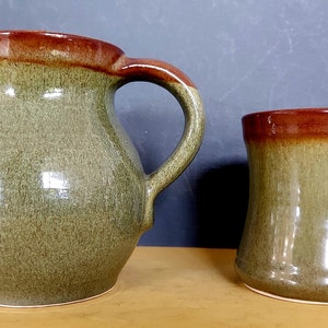 May include: A ceramic jug and mug set. The jug has a large handle and a spout. Both items are glazed in a mottled green with a brown rim. The mug has a curved handle and a similar colour scheme.