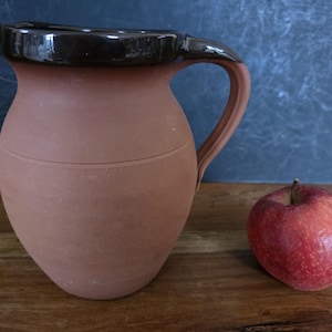 May include: A terracotta jug with a dark brown rim and handle, next to a red apple. The jug has a rounded body and a spout. The apple is red with some yellow spots. The items are on a wooden surface.