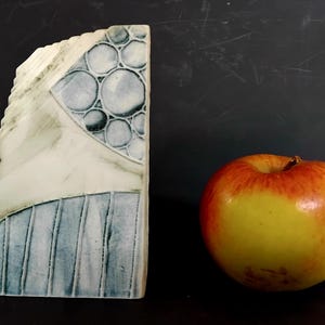 May include: A ceramic sculpture with a distinctive design. The sculpture is a mix of white, blue, and grey. It has a section with circular patterns and another with vertical lines. Beside it is a red and yellow apple. The sculpture is likely around 15-20 cm tall.