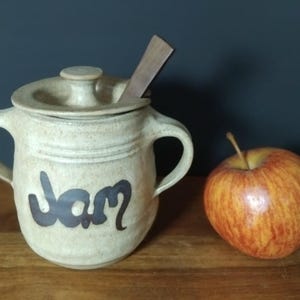 May include: A beige ceramic jam jar with a lid, two handles, and the word "Jam" in dark brown script. A wooden spoon rests in the jar. A red and yellow apple sits to the right of the jar on a wooden surface.
