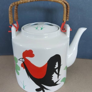 May include: White ceramic teapot with a rooster design. The teapot has a red and black rooster illustration, a woven handle, and a lid with green accents. The spout is curved, and the handle is attached with red wire.