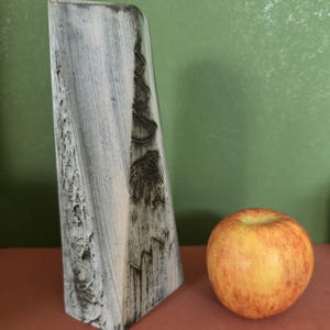 May include: A tall, angular ceramic vase with a white and grey brushed finish, featuring a textured, carved relief pattern of a pine tree and forest landscape on its side, positioned next to a single round red and yellow apple for scale.