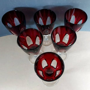 May include: A set of six ruby red glass goblets arranged in a triangular formation. Each goblet features a deep red bowl with clear vertical cut-out panels around the sides, supported by a clear glass stem and a circular base for stability.