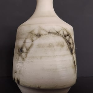 May include: Off-white ceramic vase with a narrow neck and a rounded body. The vase features a textured surface and a geometric design in dark brown. The design is a series of triangles forming an arch shape. The vase has a wide base.