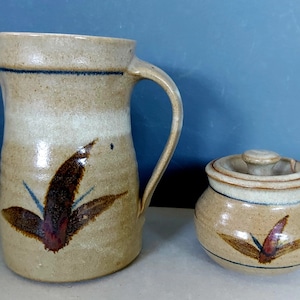 May include: A tan ceramic jug and lidded sugar bowl set. The jug has a handle and a spout. Both pieces are decorated with a dark brown and red leaf design and blue accents. The set has a rustic, handmade appearance.