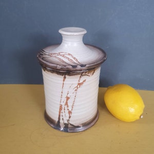 May include: A ceramic jar with a lid, cream-coloured with brown and rust accents. The jar is cylindrical with a small knob on the lid. A yellow lemon sits beside the jar. The jar's height is approximately 15 cm.