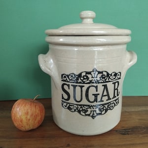May include: A cream-coloured ceramic sugar jar with a lid and handles, displaying the word "SUGAR" in black script within a decorative frame. A red and yellow apple rests beside the jar on a wooden surface.