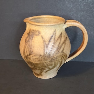 May include: A tan ceramic jug with a rooster design. The jug has a rounded body, a spout, and a curved handle. The rooster is depicted in brown lines on the front of the jug.