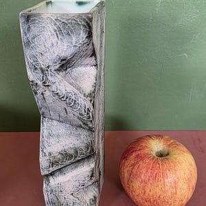 May include: A tall, rectangular ceramic vase with a textured, off-white surface featuring dark, swirling, circular patterns. The vase has a square opening with a smooth, white interior finish. A single red and yellow apple sits beside the vase.