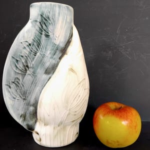 May include: A ceramic vase with an unusual, asymmetrical form. One side is light blue-grey with embossed floral patterns, the other white with similar embossed details. An apple sits beside the vase. The vase is approximately 25 cm tall.