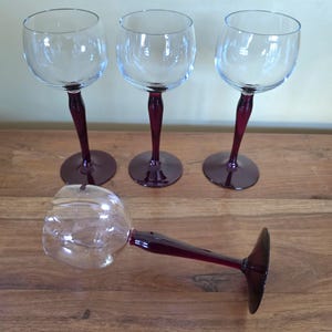 May include: Four clear wine glasses with a burgundy stem and base. Three glasses are upright, while one is lying on its side. The glasses are on a wooden surface. The stems have a twisted design.