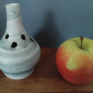 May include: A ceramic vase with a light grey and green swirl pattern and several circular openings. Next to it is a red and green apple with a stem. The objects are on a wooden surface.