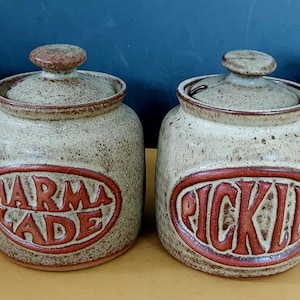 May include: Four ceramic storage jars with lids, labelled "Apple Sauce", "Marmalade", "Pickles", and "Jam". The jars are beige with brown lettering and accents, ideal for the kitchen.