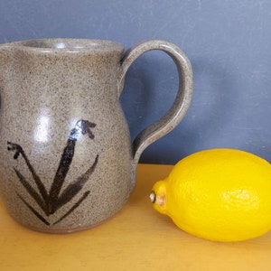 May include: A speckled stoneware jug with a curved handle and a spout. The jug has a dark brown floral design on the front. A bright yellow lemon sits next to the jug. The background is a muted blue and yellow.