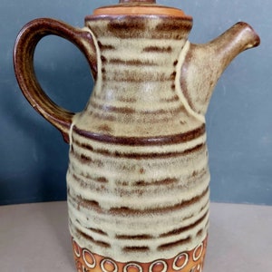 May include: A ceramic pitcher with a brown handle, spout, and lid. The body is beige with horizontal brown stripes. The base features a decorative pattern of circles and geometric shapes in shades of brown and orange. The pitcher is designed for serving beverages.