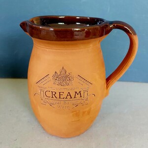 May include: A terracotta-coloured ceramic jug with a dark brown rim and handle. The jug features a spout and a printed label with the word "Cream" and "Royal Barum Ware". Likely used for serving cream or other liquids, it is a classic design.