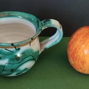 May include: A handcrafted ceramic creamer with a green and white pattern, a handle, and a spout, beside a red and yellow apple. The creamer has a unique, artistic look. The apple has a brown stalk.