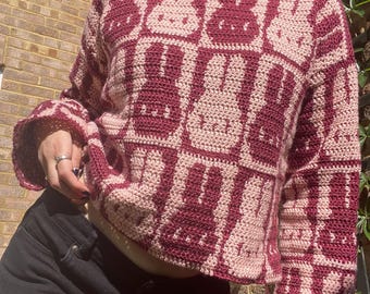May include: A pink and burgundy crocheted jumper with a repeating pattern of bunny rabbits. The jumper has long sleeves and a relaxed fit.