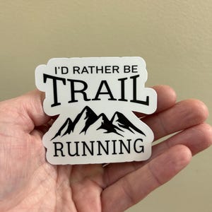 May include: White sticker with black text that reads "I'D RATHER BE TRAIL RUNNING." The sticker features a mountain range graphic. The sticker is held in a hand.