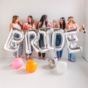 May include: Five friends pose in front of a silver foil 'PRIDE' balloon arch. They are all wearing casual clothing and holding drinks.