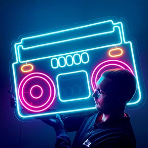 K&ouml;nnte beinhalten: Ein Neon-Schild in Form einer Vintage-Boombox mit blauen, pinken und gelben Lichtern. Das Schild wird von einer Person gehalten, die ein schwarzes Hemd und eine Brille tr&auml;gt.