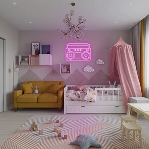K&ouml;nnte beinhalten: Ein pinkfarbenes Neon-Boombox-Schild an einer pinkfarbenen Wand mit einem wei&szlig;en Kinderbett und einer gelben Couch im Hintergrund.