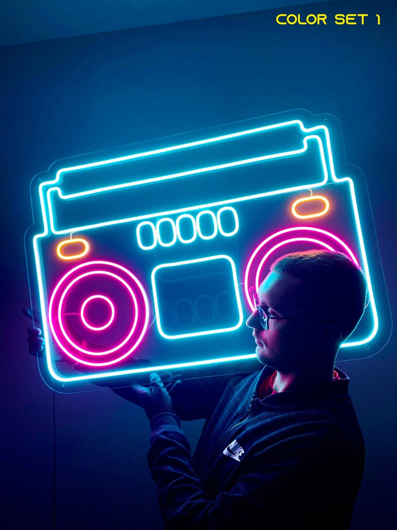 K&ouml;nnte beinhalten: Ein Neon-Schild in Form einer Vintage-Boombox mit blauen, pinken und gelben Lichtern. Das Schild wird von einer Person gehalten, die ein schwarzes Hemd und eine Brille tr&auml;gt.