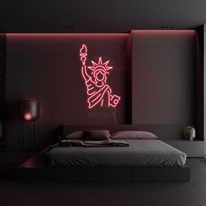 May include: Pink neon sign of the Statue of Liberty with the word "NEONIX" below it, hanging on a black wall above a black bed with gray bedding.