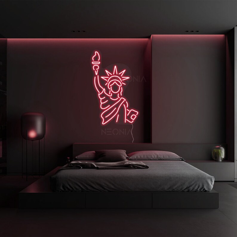 May include: Pink neon sign of the Statue of Liberty with the word "NEONIX" below it, hanging on a black wall above a black bed with gray bedding.