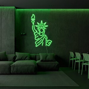 May include: Green neon sign depicting the Statue of Liberty, with a torch in her right hand, on a dark green wall in a living room setting.