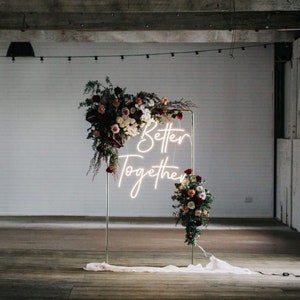 May include: A gold metal frame with a white neon sign that reads "Better Together" surrounded by burgundy and pink flowers.