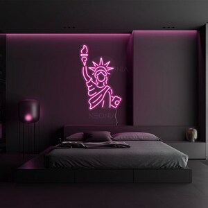 May include: Pink neon sign of the Statue of Liberty in a dark bedroom with a black bed and a black lamp. The sign says "NEONIX" at the bottom.